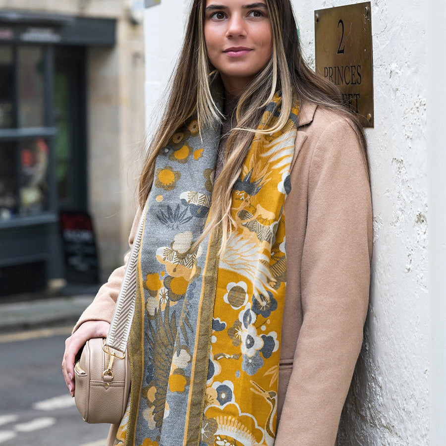 Woman with long hair wearing a beige coat and a floral jacquard scarf stands beside a wall featuring a brass plaque that reads 2 Princes Street, holding a beige handbag.
