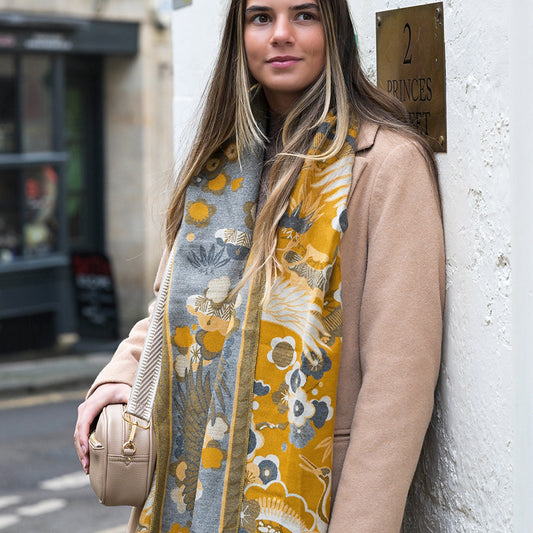 Woman with long hair wearing a beige coat and a floral jacquard scarf stands beside a wall featuring a brass plaque that reads 2 Princes Street, holding a beige handbag.