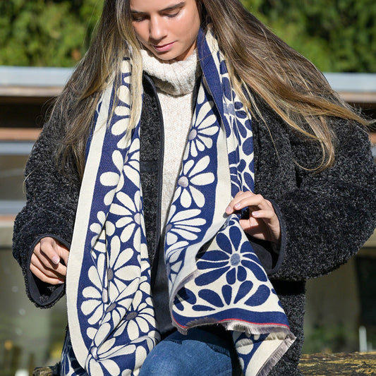 A person dressed in a thick black jacket, cream jumper, and blue jeans sits outdoors, holding a large navy retro daisy scarf adorned with a charming floral pattern.