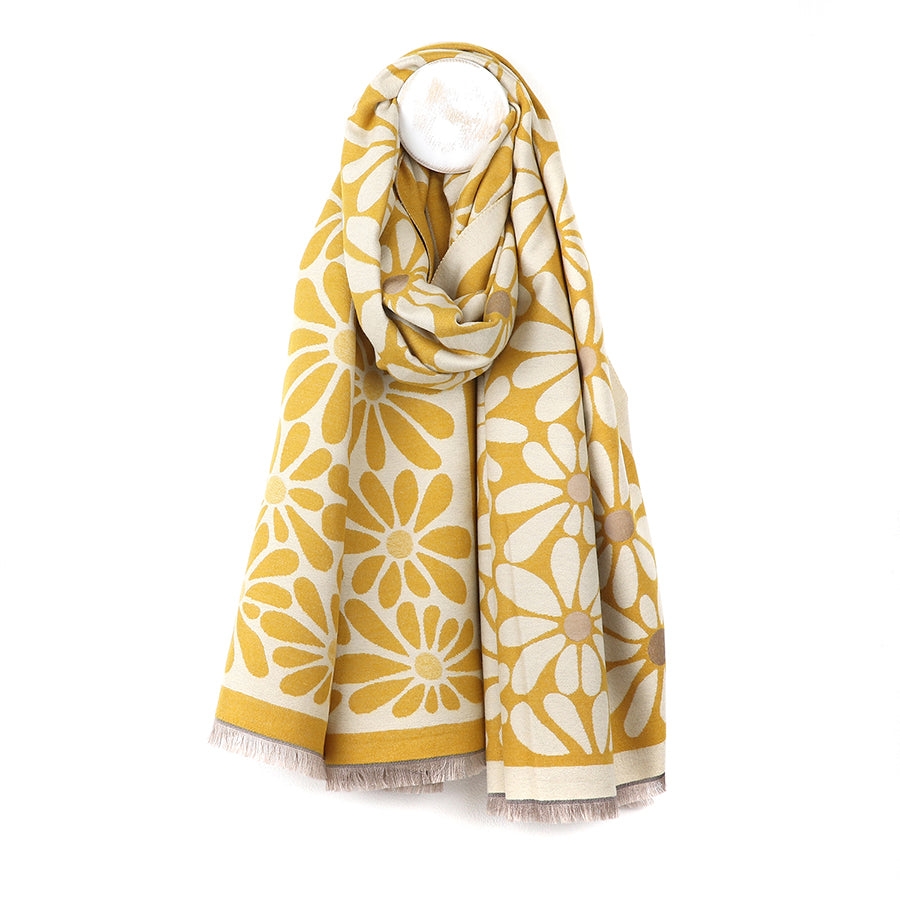 A yellow and white Jacquard scarf featuring a large daisy floral pattern hangs on a white hook, its fringed ends lending charm to this cheerful, vintage-inspired accessory.