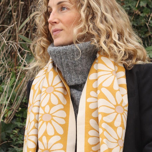 A woman with curly blonde hair wears a grey polo neck jumper, black coat, and a yellow retro daisy scarf, standing outdoors by green foliage.