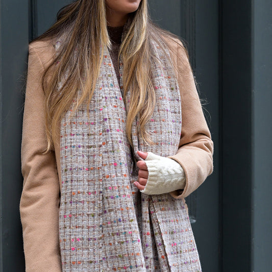 A woman in a beige coat, cream fingerless gloves, and a cosy winter accessory—a grey woven-texture scarf with colourful specks—stands against a dark-coloured door.