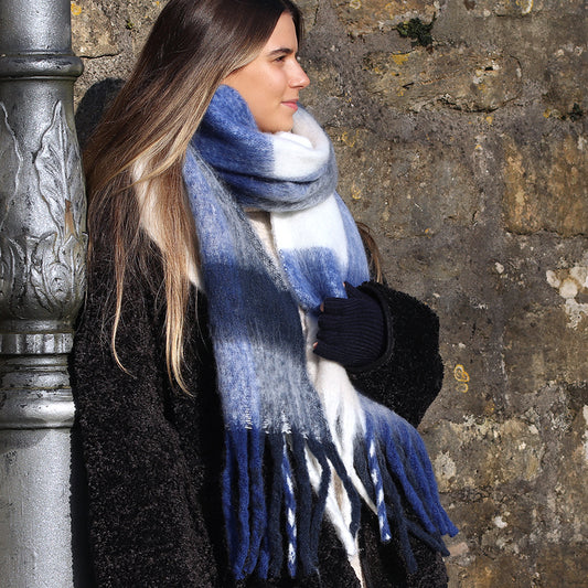 Woman with long hair wearing a cosy winter accessory—a thick blue and white striped scarf with a soft texture—and black coat stands outdoors beside a stone wall.