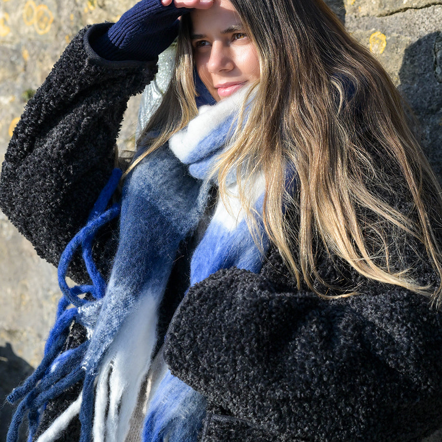 A person in a thick coat and grey woven scarf stands outdoors by a stone wall, shielding their eyes from the sunlight with one hand.