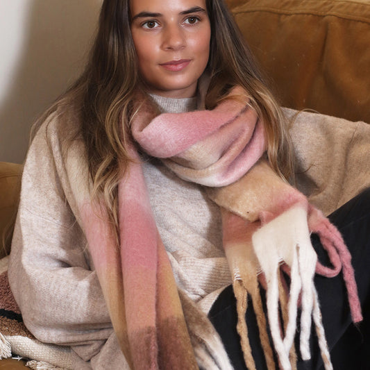 A woman with long brown hair sits on a sofa wearing a beige jumper and a large, pale pink chunky checked scarf with fringe.