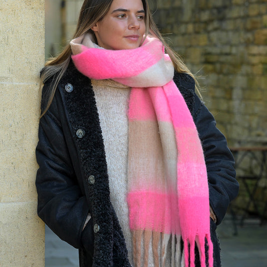 A woman in a dark coat and thick checked scarf stands outdoors against a stone wall, gazing to the side.