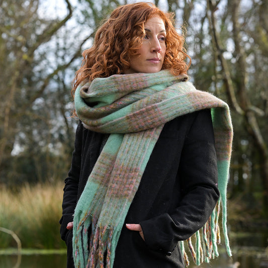 A woman with curly red hair stands outdoors in a black coat, wrapped in a chunky checked scarf—a perfect winter accessory—among trees and grass.