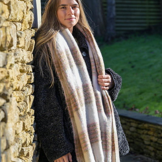 A woman in a dark coat and beige chunky checked scarf stands beside a stone wall outdoors on a sunny day, her cosy accessory adding warmth and charm to her appearance.