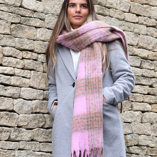 A person in a grey coat and a chunky checked scarf stands before a stone wall, hands tucked into their coat pockets.