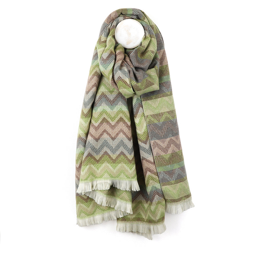 A vibrant chevron scarf in shades of green, beige, and brown with fringed edges is draped over a white hook.