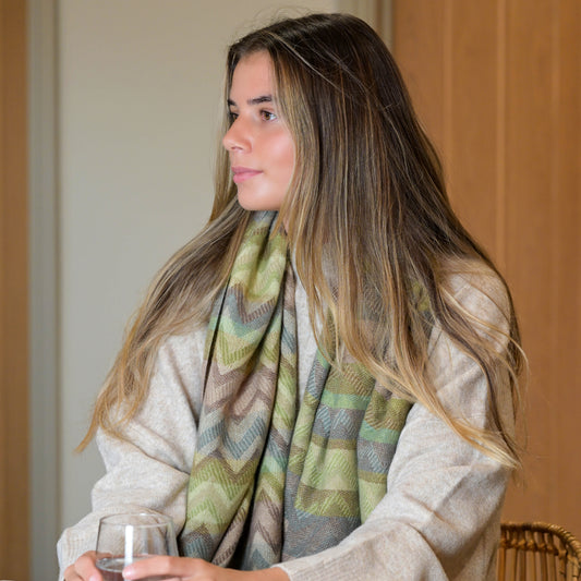 A woman with long, straight hair sits indoors, wearing a light jumper and a colourful scarf, holding a glass of water.