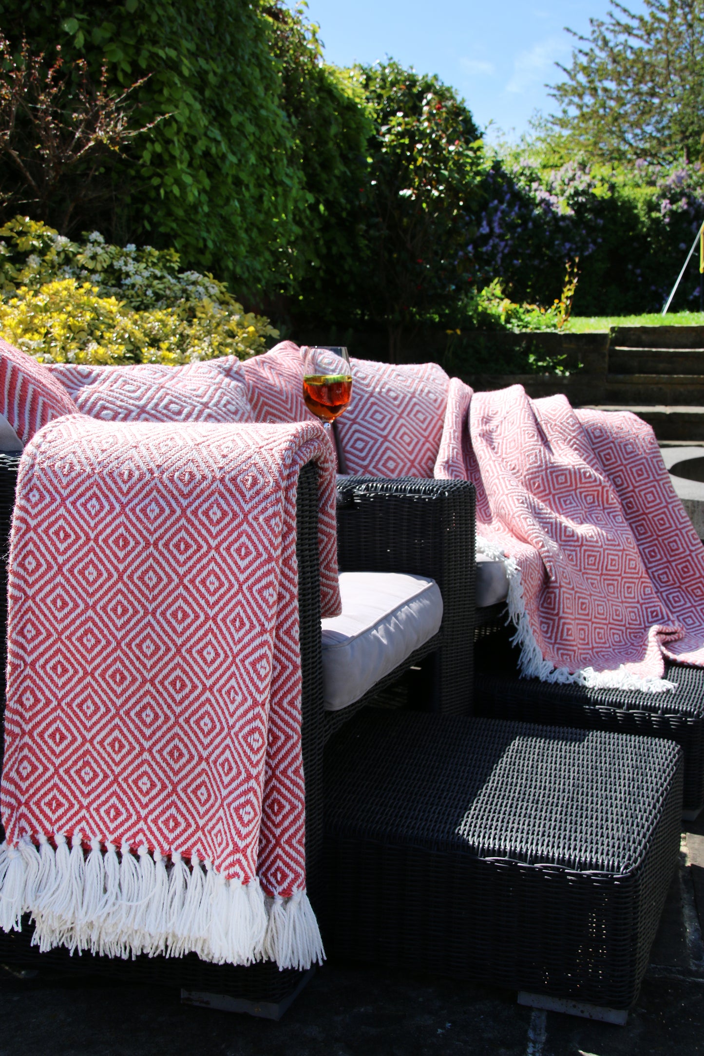 Outdoor patio chairs with UV-stable colour in red and white diamond-patterned throws, crafted from recycled plastic bottles, and a glass of rosé on the armrest, set on a sunny day in the garden.