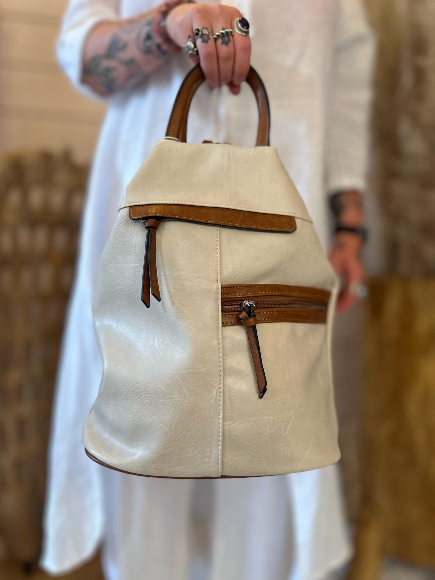 A person dressed in white holds a cream-coloured faux leather rucksack with brown accents and a front zip pocket, shown in focus against a blurred background.