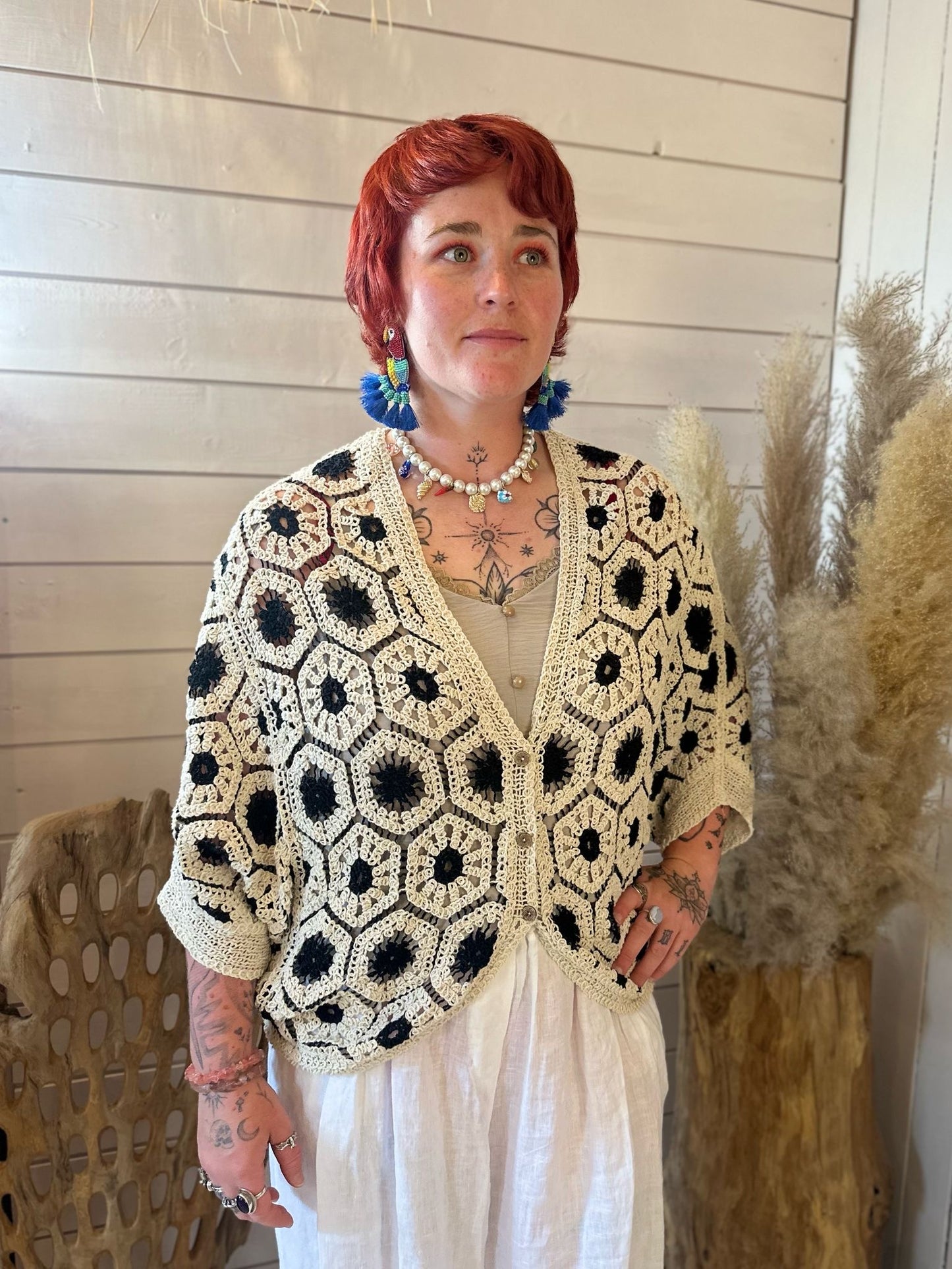 A person with short red hair and tattoos wears a black crochet cardigan featuring black and white hexagon patterns, a white skirt, and blue flower earrings—showcasing a versatile wardrobe beside decorative pampas grass indoors.