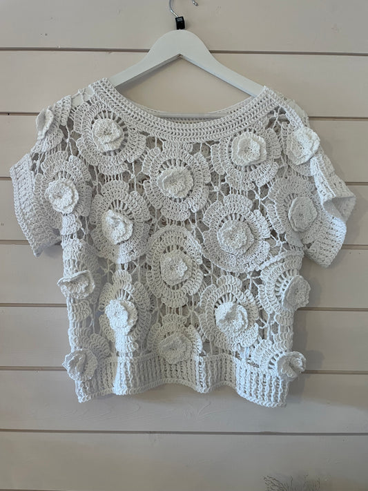 A white floral jumper with a crochet short-sleeve design and flower pattern hangs on a white hanger against a light wooden wall—ideal for layering in spring.
