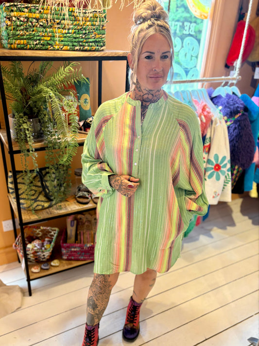 A woman with tattoos stands indoors on a light wooden floor, wearing a fashionable piece—a Green Stripe Shimmer Shirt Dress—and purple lace-up boots. Shelves with plants and décor are behind her.