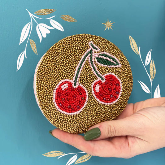 A hand with green nail varnish holds a cherry coin purse adorned with two red cherries and green leaves, set against a teal background with white and gold leaf designs—ideal as a vegan accessory or unique Christmas present.