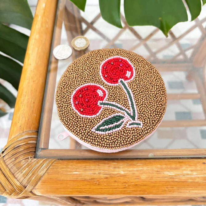A cherry coin purse adorned with red and green beaded cherries rests on a glass table beside two coins and green leaves—an adorable vegan accessory and an ideal Christmas gift.