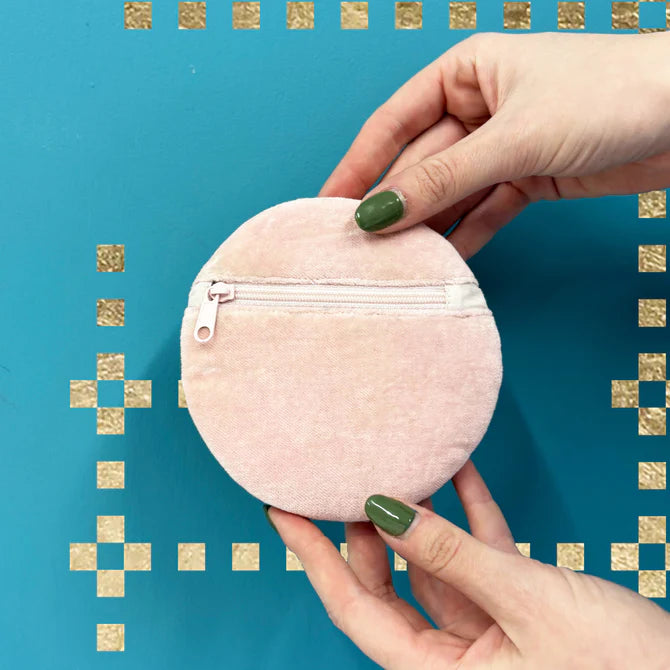 Two hands with green nail varnish hold a round, light pink cherry coin purse—a fashionable vegan accessory—in front of a blue wall adorned with gold square decorations.