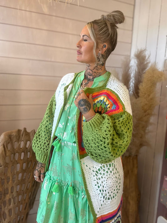 A woman with tattoos is wearing a green dress and a handcrafted crochet long cardigan with multicoloured patterns, standing indoors next to a decorative chair and dried plants.