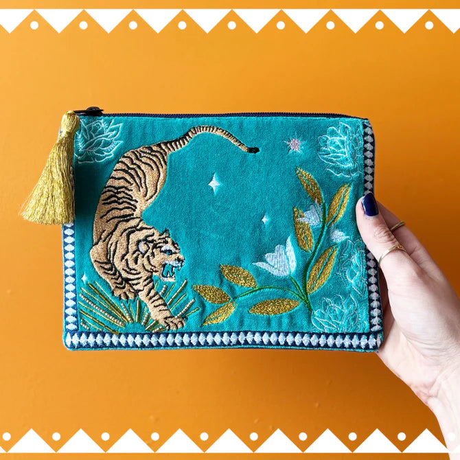 A hand holds a teal embroidered Tiger Makeup Bag featuring a tiger, flowers, leaves, and a gold tassel—vegan-friendly and ideal as a unique Christmas present—set against an orange background.