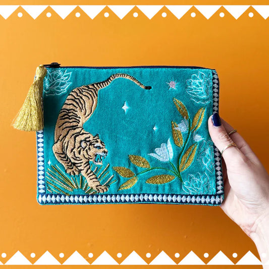 A hand holds a teal embroidered Tiger Makeup Bag featuring a tiger, flowers, leaves, and a gold tassel—vegan-friendly and ideal as a unique Christmas present—set against an orange background.