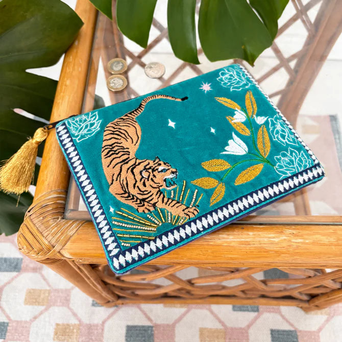 A teal Tiger Make-up Bag featuring an embroidered tiger and floral pattern, a gold tassel, and coins set nearby on a glass-topped wicker table with green leaves above—ideal as a Christmas present.