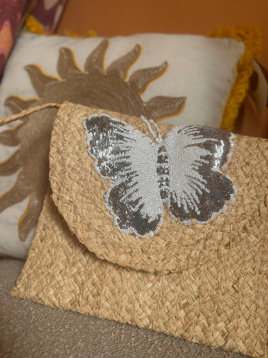 A woven straw bag with a silver and white sequin butterfly on the flap, this stylish handbag sits in front of a decorative cushion featuring a sun design.