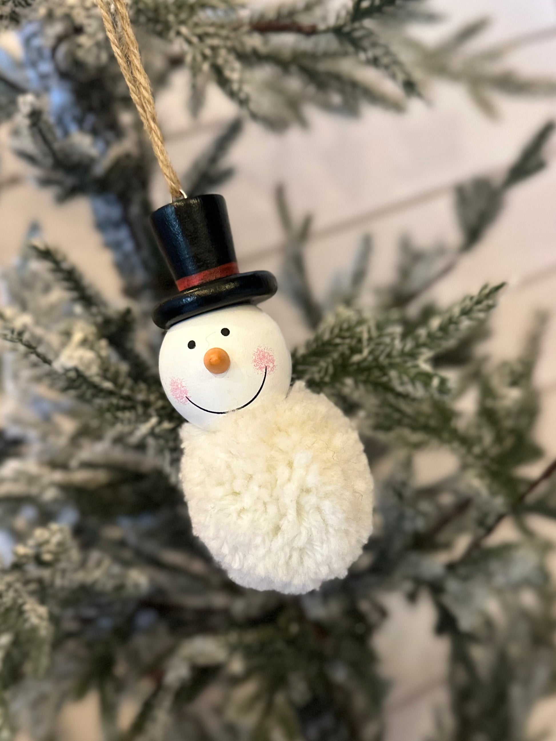 A snowman decoration with a black top hat and a pom-pom body hangs from a snowy pine branch, bringing festive charm to your holiday décor.