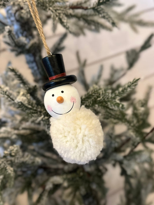 A snowman decoration with a black top hat and a pom-pom body hangs from a snowy pine branch, bringing festive charm to your holiday décor.