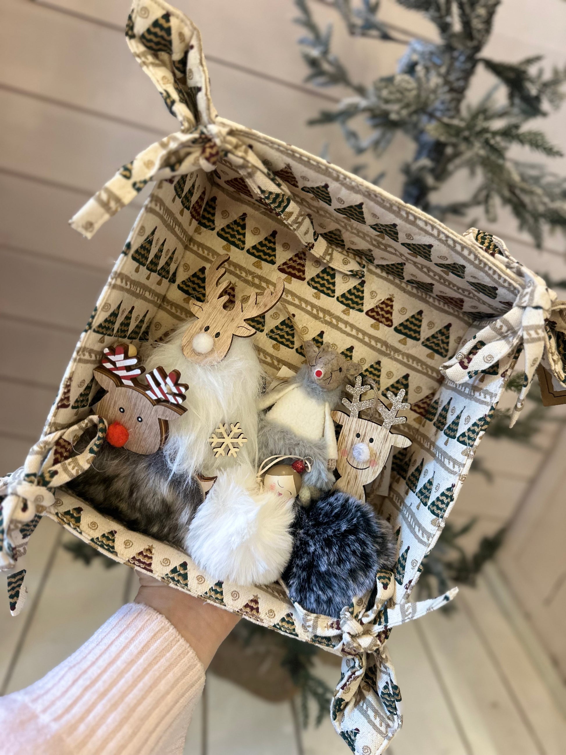 A hand holds a fabric basket with a Christmas tree pattern, filled with six festive animal decorations including a Fluffy Hanging Reindeer, in front of a decorated tree—ideal for festive décor or Christmas tree decoration.