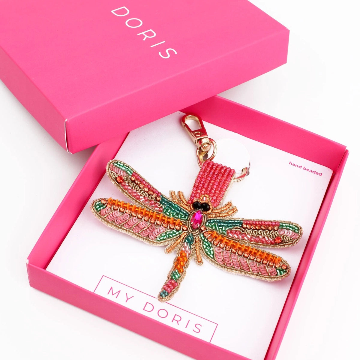 The pink box reveals a handcrafted dragonfly keyring adorned with intricate beadwork, featuring multicoloured wings and a pink gem. The open lid proudly displays the brand name Doris.