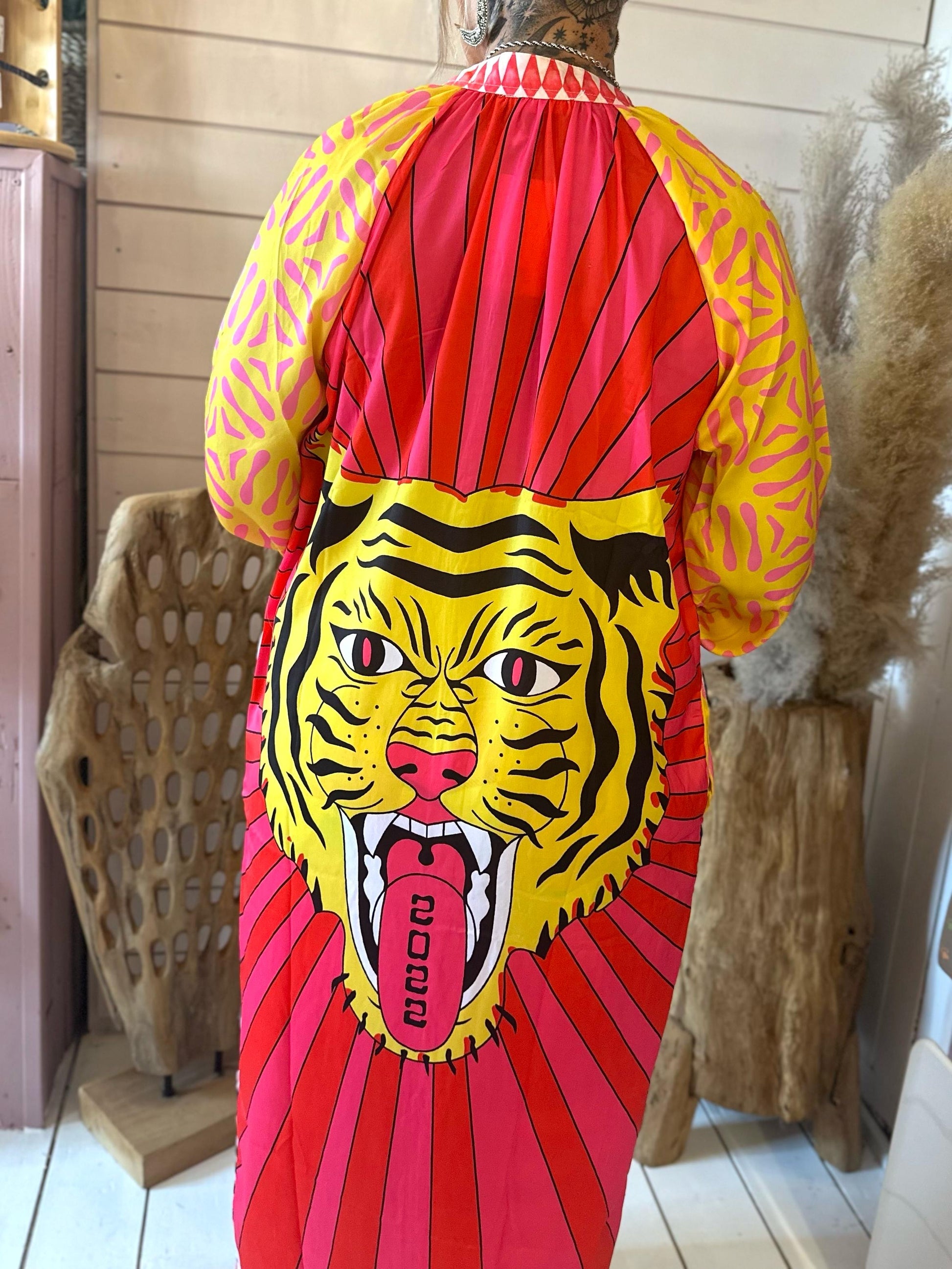 Person facing away, wearing a colourful robe featuring a large graphic of a tigers face with the year 2022 on its tongue; the background includes textured decorative items and wooden panelling.