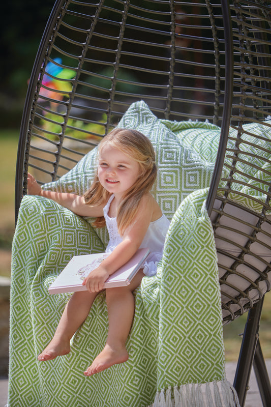 A young girl sits in a hanging chair with a green patterned sustainable throw, made from recycled plastic bottles, holding an open book and smiling outside.