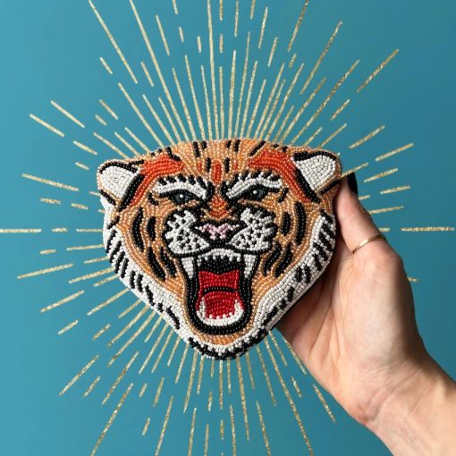 A hand holds a vegan-friendly beaded accessory in the shape of a roaring tiger’s head, set against a blue background adorned with gold radiating lines.