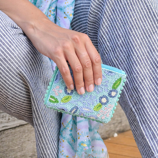 A hand cradles a small handbag, adorned with floral embellishments and aqua blue beading, resting on a lap draped in striped fabric clothing.