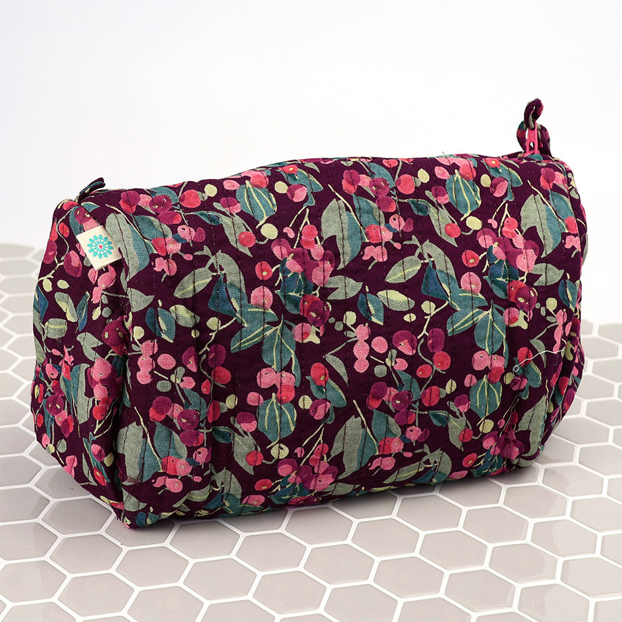 The Winter Berry Quilted Make-up Bag boasts a maroon background with pink and green floral patterns, ideal for keeping your beauty essentials on a chic white hexagonal tiled surface.