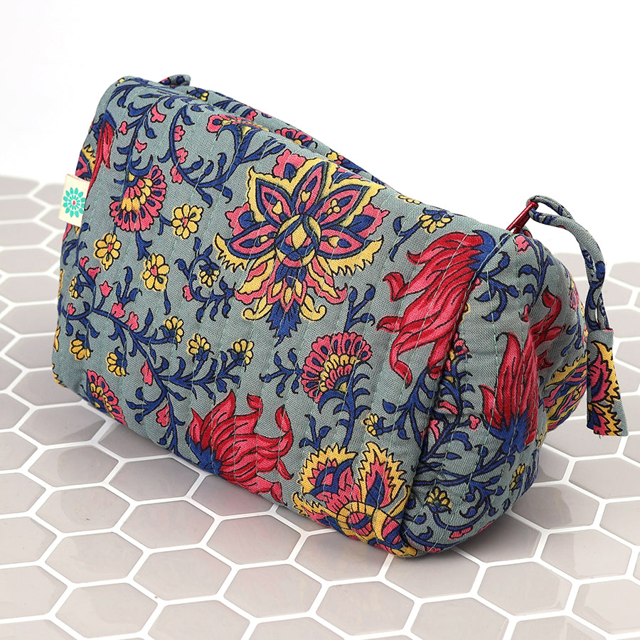 A rectangular quilted make-up bag with a colourful floral pattern in red, yellow and blue on a grey background, resting on a hexagonal tiled surface.