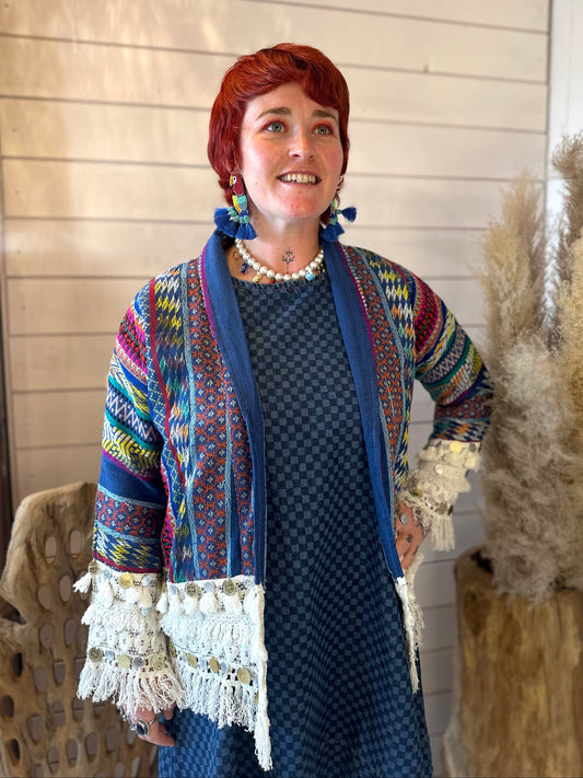 A woman with short red hair is wearing a colourful, patterned jacket over a blue dress, standing indoors next to pampas grass and wood-panelled walls.