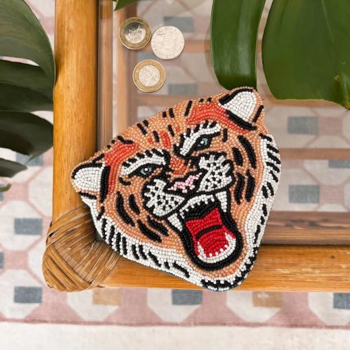 A vegan-friendly Tiger Coin Purse, shaped like a roaring tiger’s face and decorated with beads, rests on a wooden table beside three coins and some green leaves.
