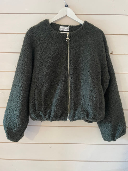 A forest green, zip-up, textured fleece casual jacket with long sleeves, hanging on a white hanger against a light wooden wall; this vegan-friendly piece is ideal for cosy style.
