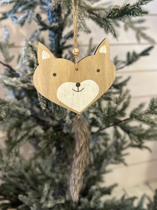 A vegan-friendly wooden fox Christmas decoration with a faux fur tail hangs from a string on a Christmas tree adorned with green and frosted branches.
