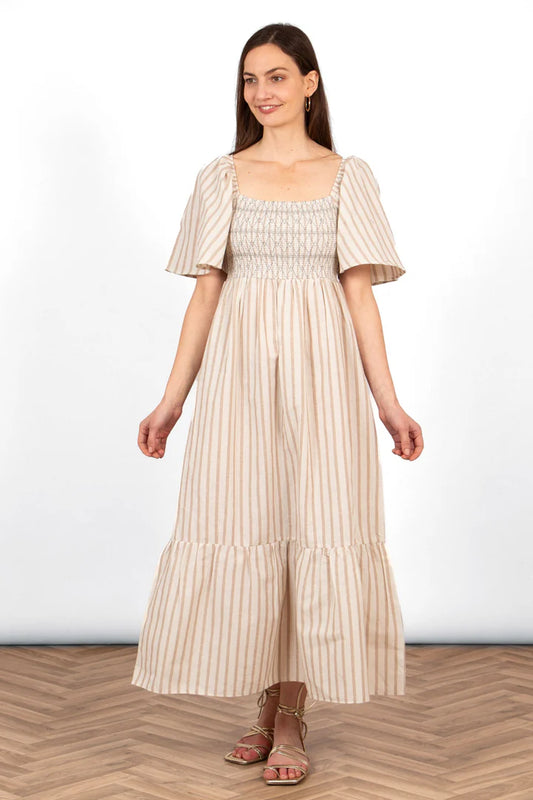 A woman exudes elegance in a striped, short-sleeved Briar Milkmaid Dress as she stands on a wooden floor, facing forward with a neutral expression, embodying the poise of a true fashion enthusiast.