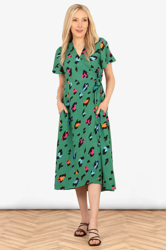 Woman in an exclusive green wrap dress with colourful abstract patterns, reminiscent of the luxurious Addie Wrap Large Leopard Print Dress, stands with hands in pockets on a wooden floor.