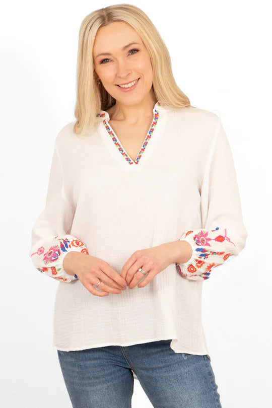 A person with long blonde hair wearing an elegant blouse featuring colourful floral embroidery on the cuffs and neckline, paired with blue jeans, standing and smiling against a white background.