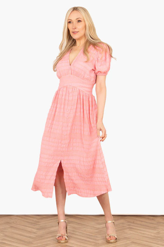 A woman in a coral, ankle-length Oliva Button Down Tea Dress with a V-neck and puff sleeves stands elegantly on a wooden herringbone floor, facing forwards.