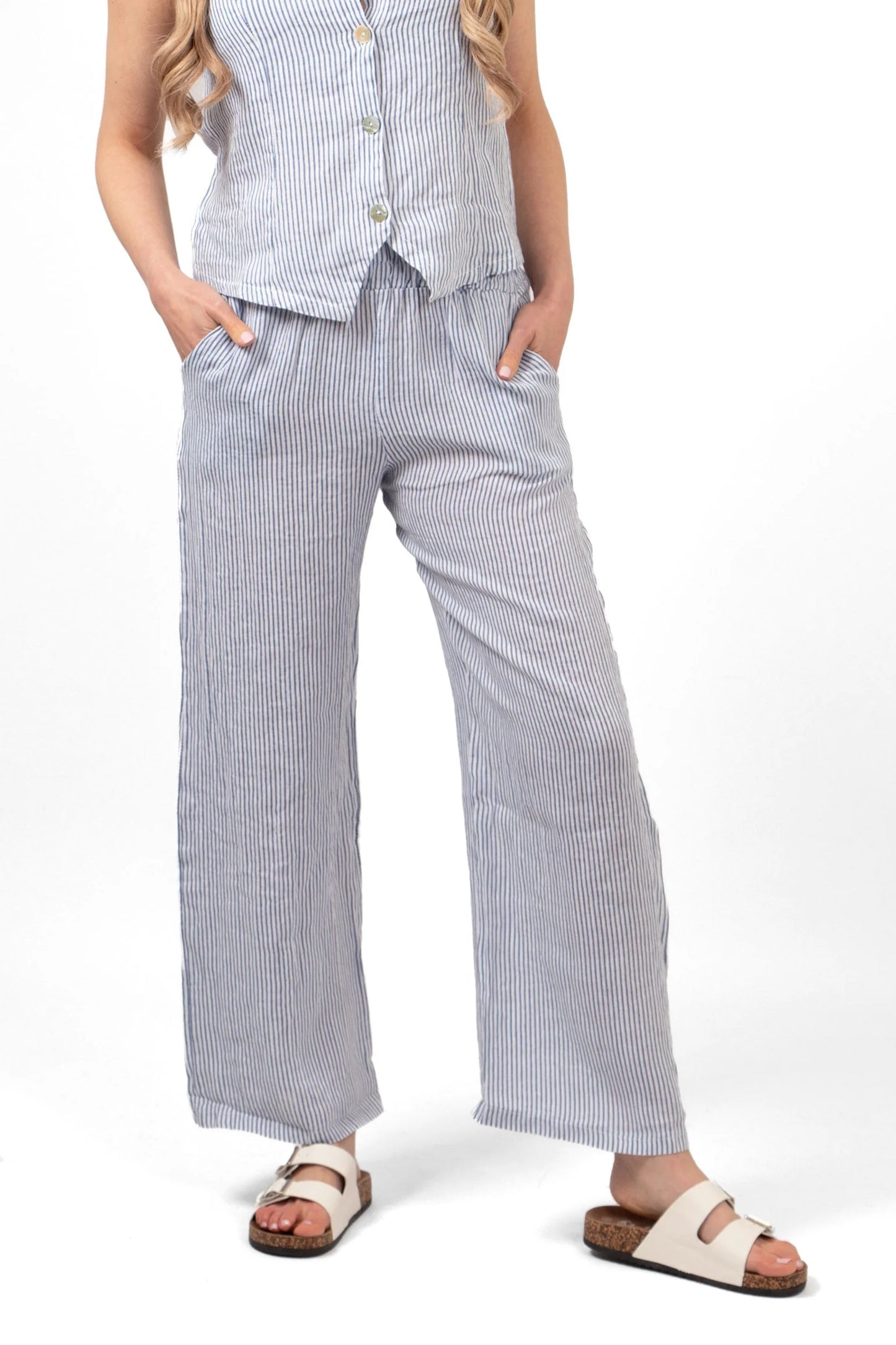 A person is wearing a sleeveless, buttoned, pinstriped Italian linen top and matching wide-leg trousers with an elasticated waist, hands in pockets, paired with white slide sandals. The head is not visible.
