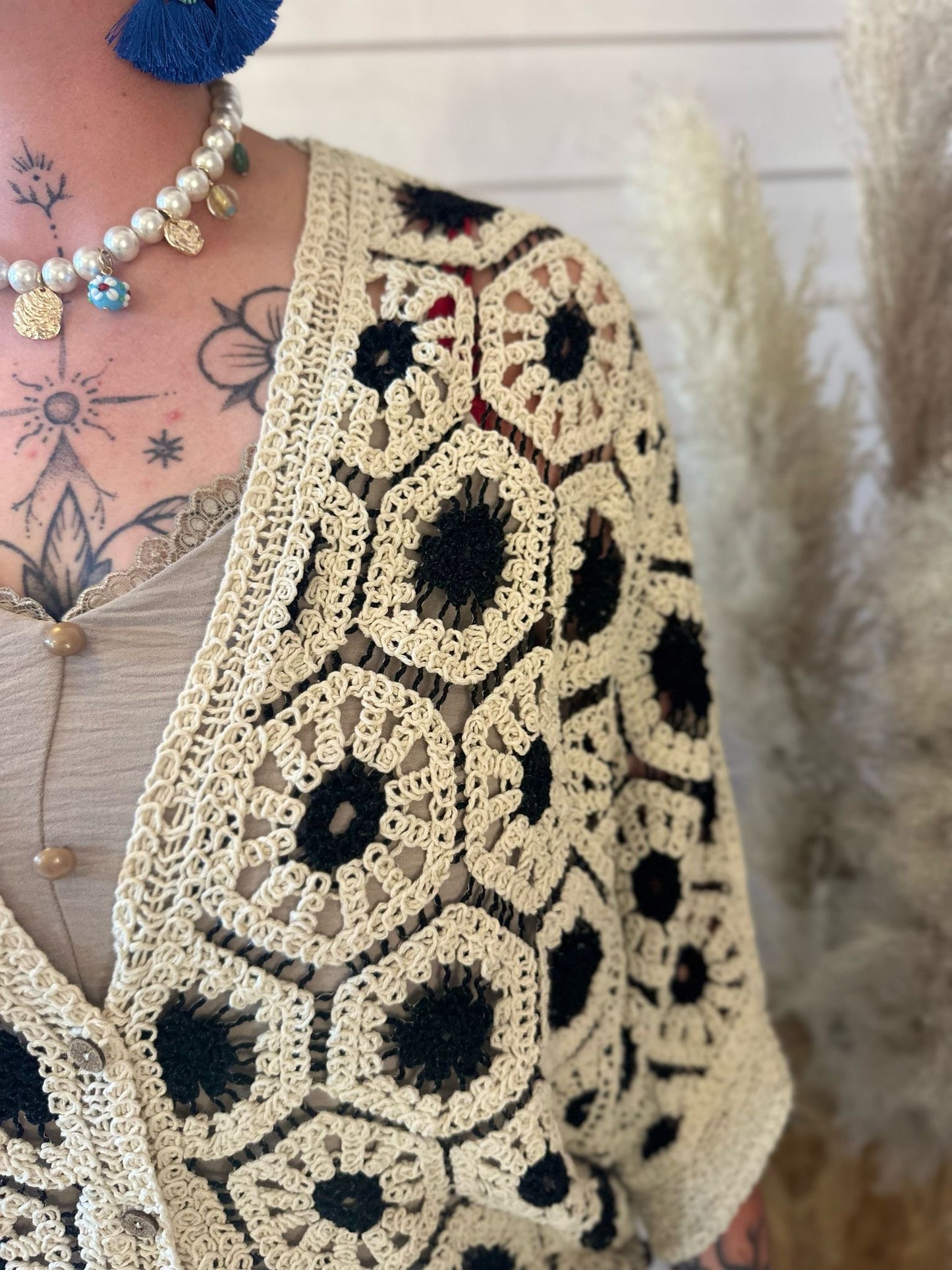 A person is wearing a black crochet cardigan with diamond detailing, a beige buttoned top, a pearl necklace, blue tassel earrings, and displaying chest tattoos. Pampas grass adds to the versatile wardrobe backdrop.