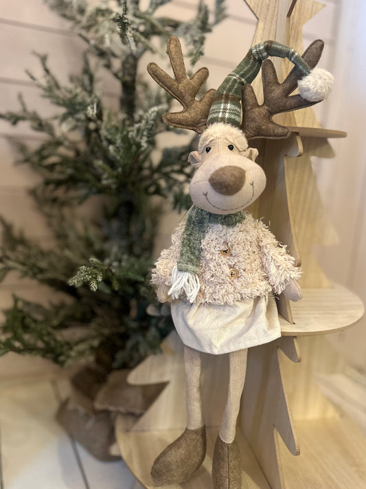 A plush Dangly Legs Christmas Deer Decoration wearing a green checked hat and scarf sits atop a wooden tree, with a small artificial pine tree providing festive holiday décor in the background.