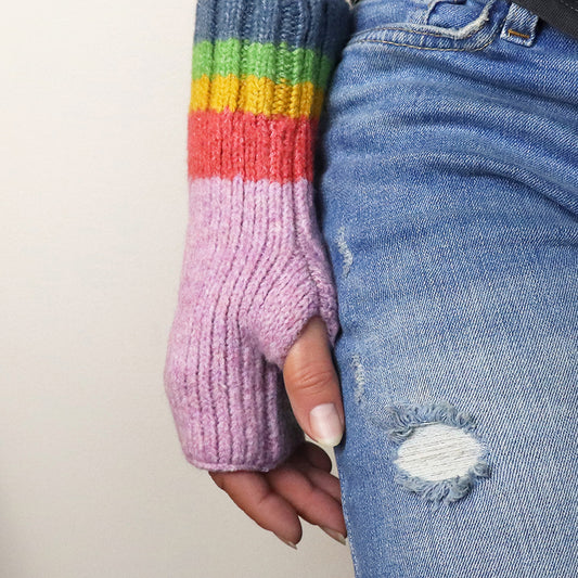 A person wearing distressed blue jeans and knitted, cruelty-free mauve rainbow mittens with fingerless cuffs.